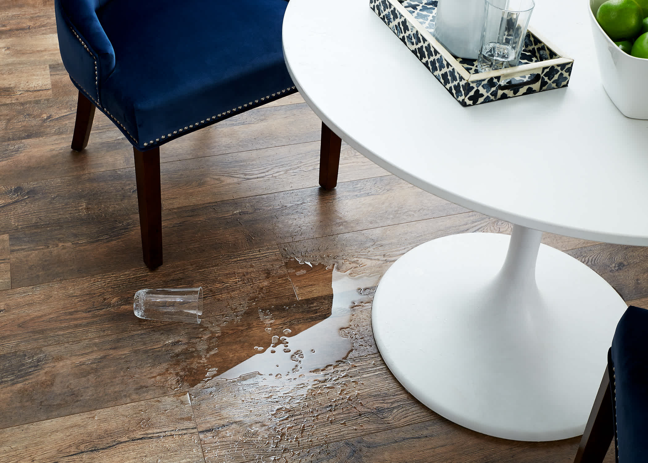 dark brown water-resistant laminate floor with spilled glass of water on floor in front of dark blue velvet chair and white oval table