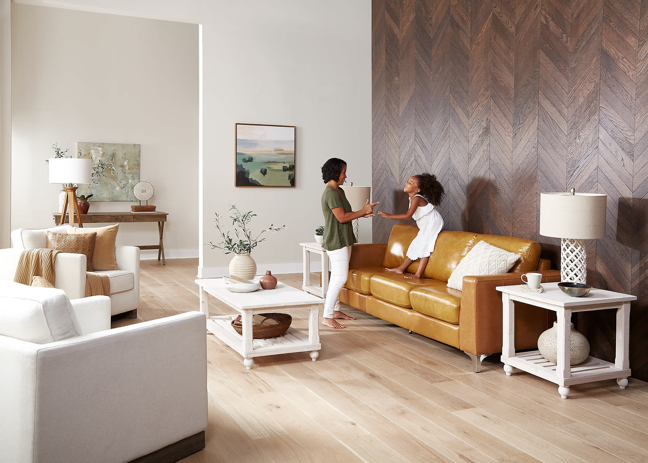 blonde hardwood floors in living room plus dark brown chevron hardwood floor on accent wall behind caramel leather sofa with woman in green shirt playing with toddler in white dress standing on the sofa