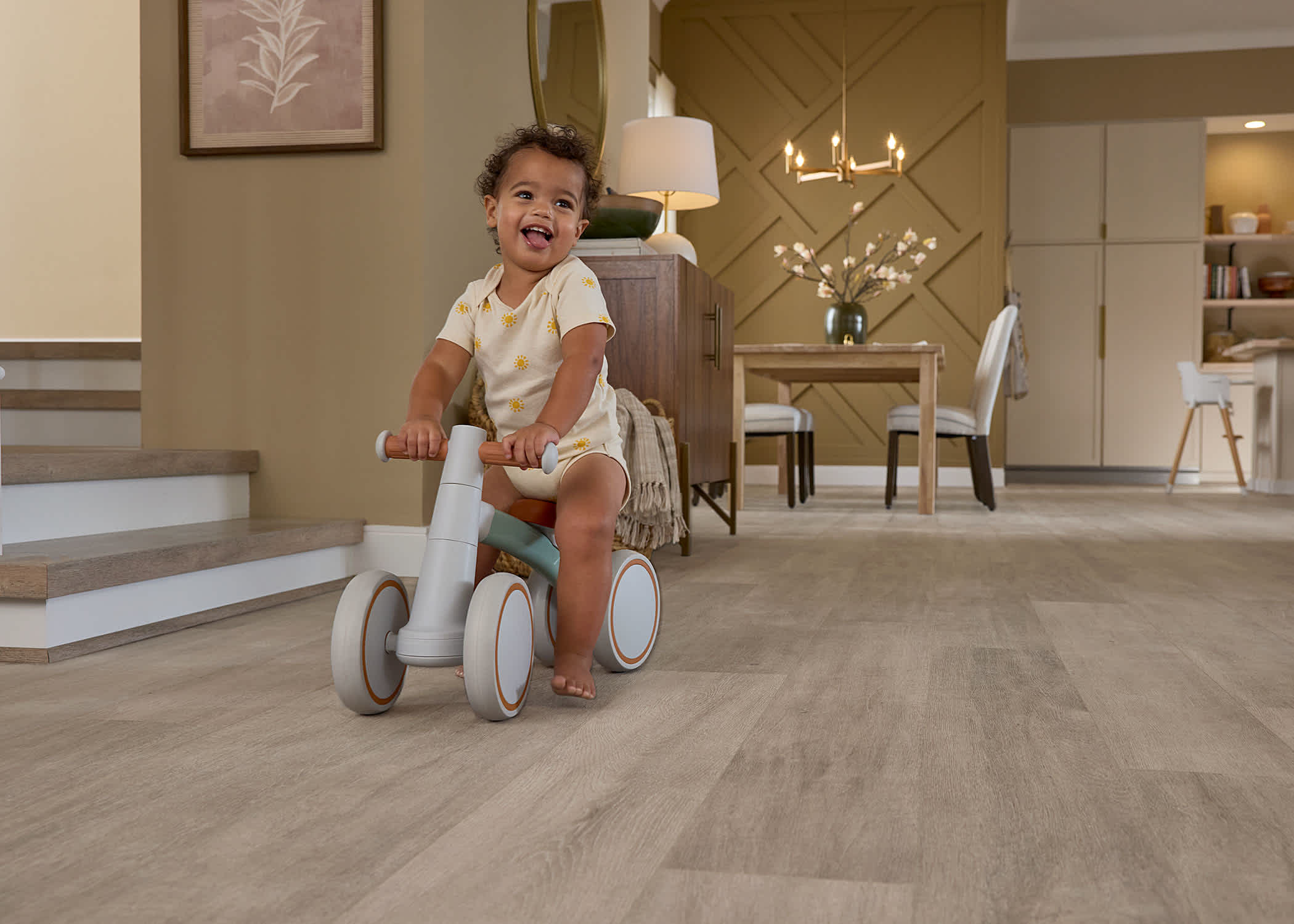 beige waterproof rigid vinyl plank floor in open concept living and dining room with baby riding on tricycle next to stairs with dining area in background