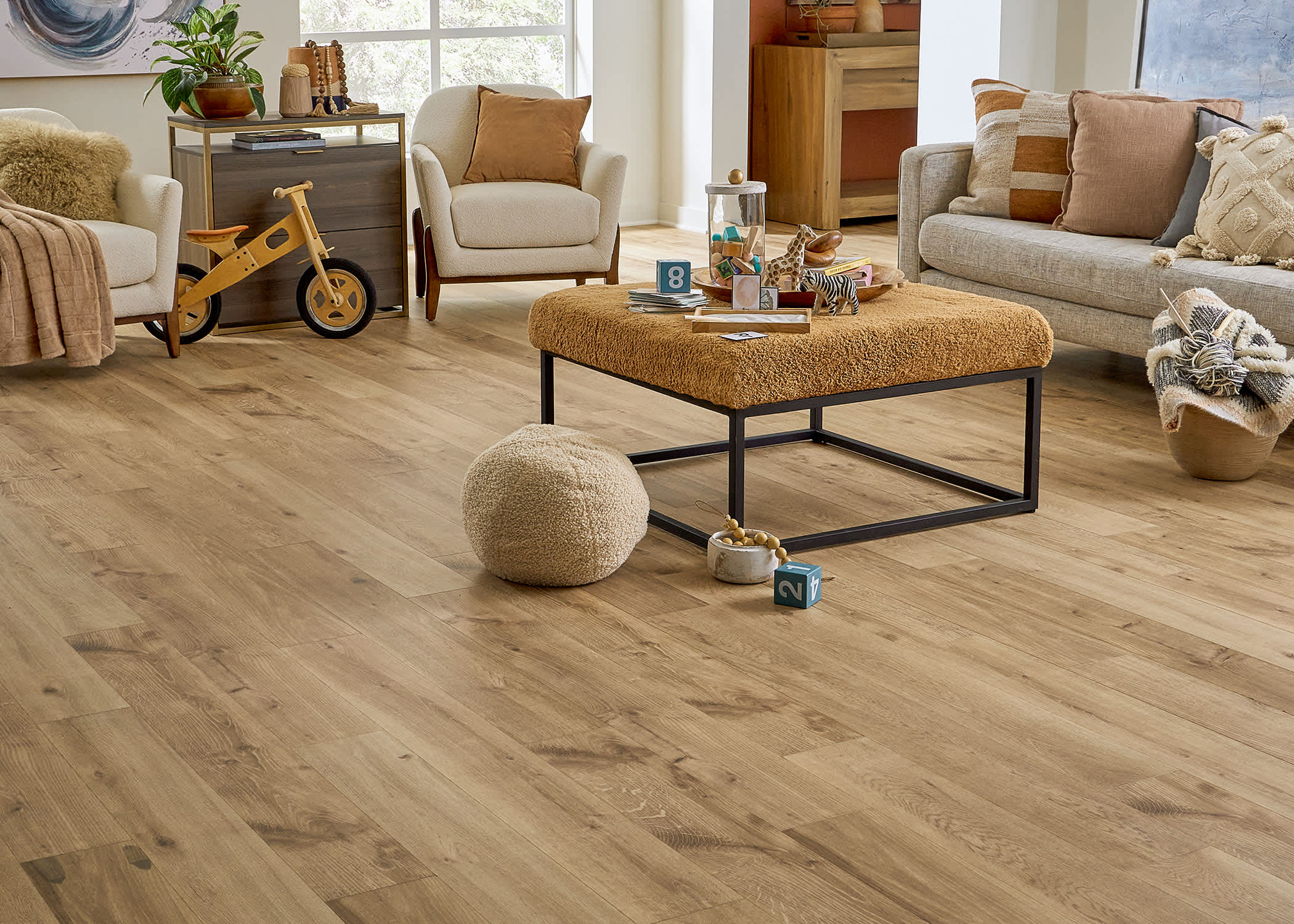 5mm with Pad Yorkshire Oak Waterproof Rigid Vinyl Plank Flooring in living room with beige furnishings plus gold upholstered ottoman and wooden tricycle and knotting material in a basket