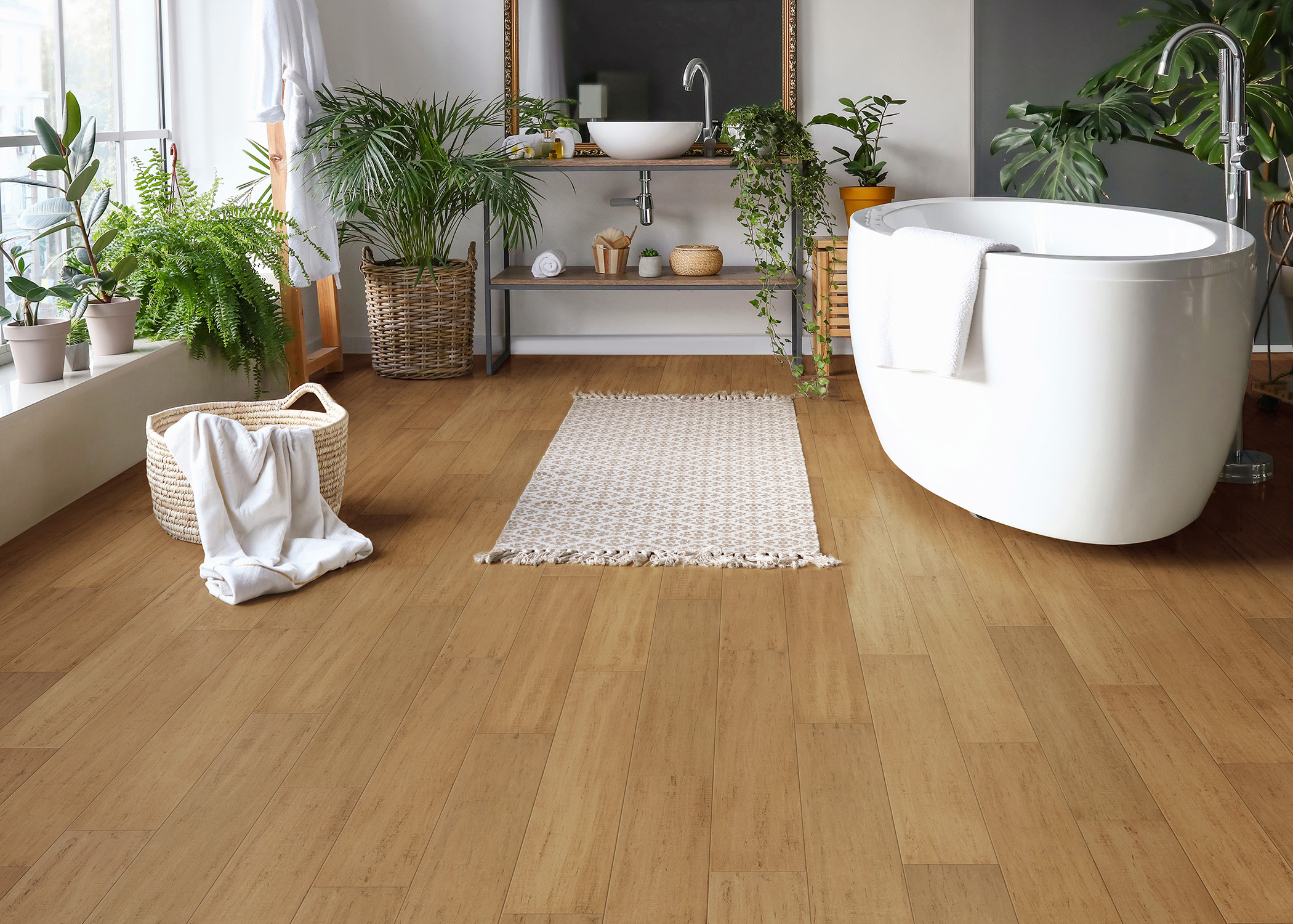 blonde water resistant engineered bamboo floor in bathroom with freestanding oval bathtub plus window sill filled with green plants and small off white bath mat on floor