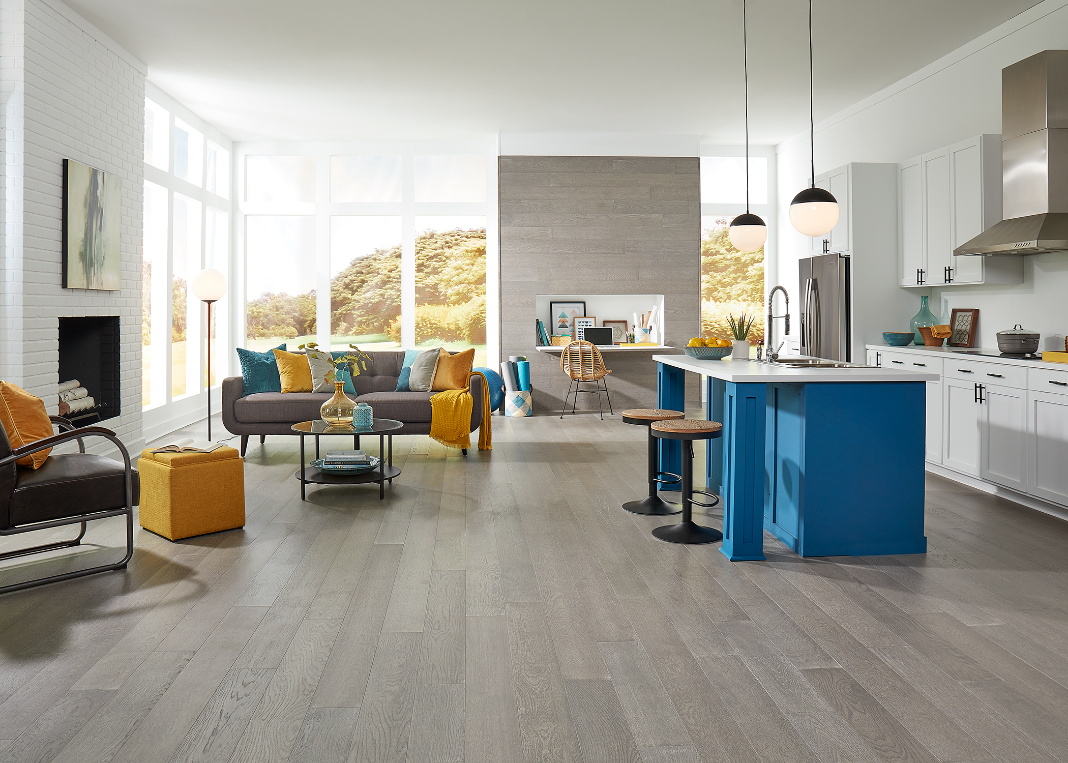 gray engineered hardwood floor in open concept living room and kitchen with blue island with white countertops plus wood accent wall with built in desk and dark gray sofa with yellow and blue accent pillows