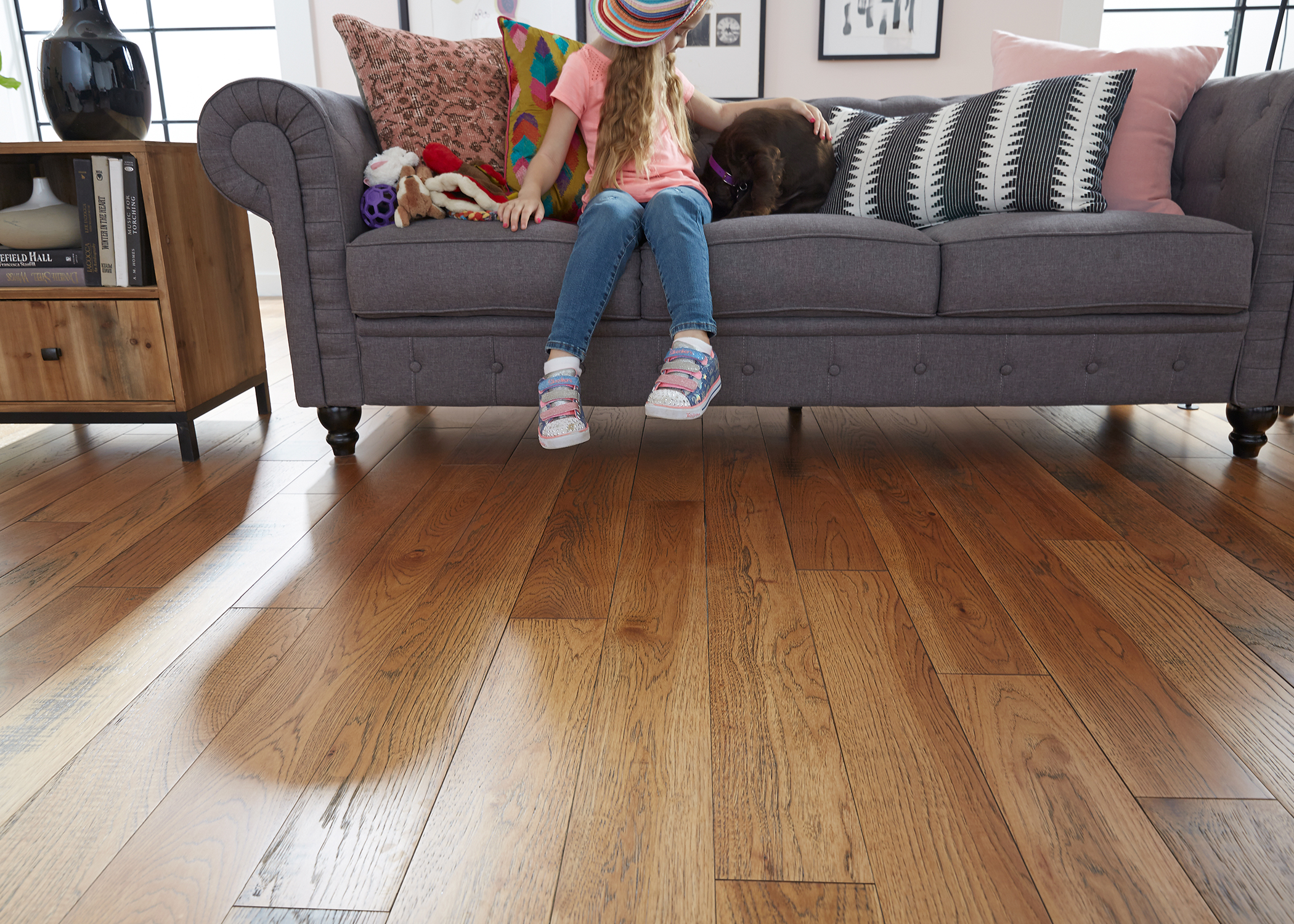 medium brown solid hardwood floor in living room with young child and dog sitting on gray sofa with pink and black accent pillows