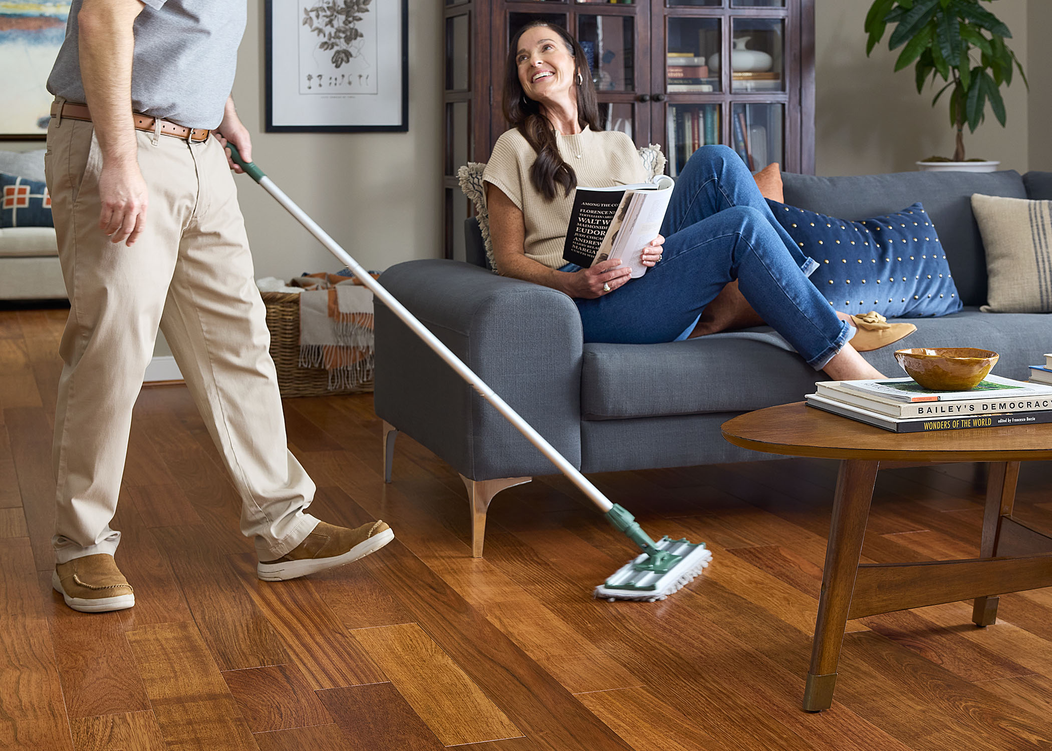 person cleaning reddish brown solid hardwood floor while woman laughing lifts her feet while sitting on a dark gray sofa reading a magazine