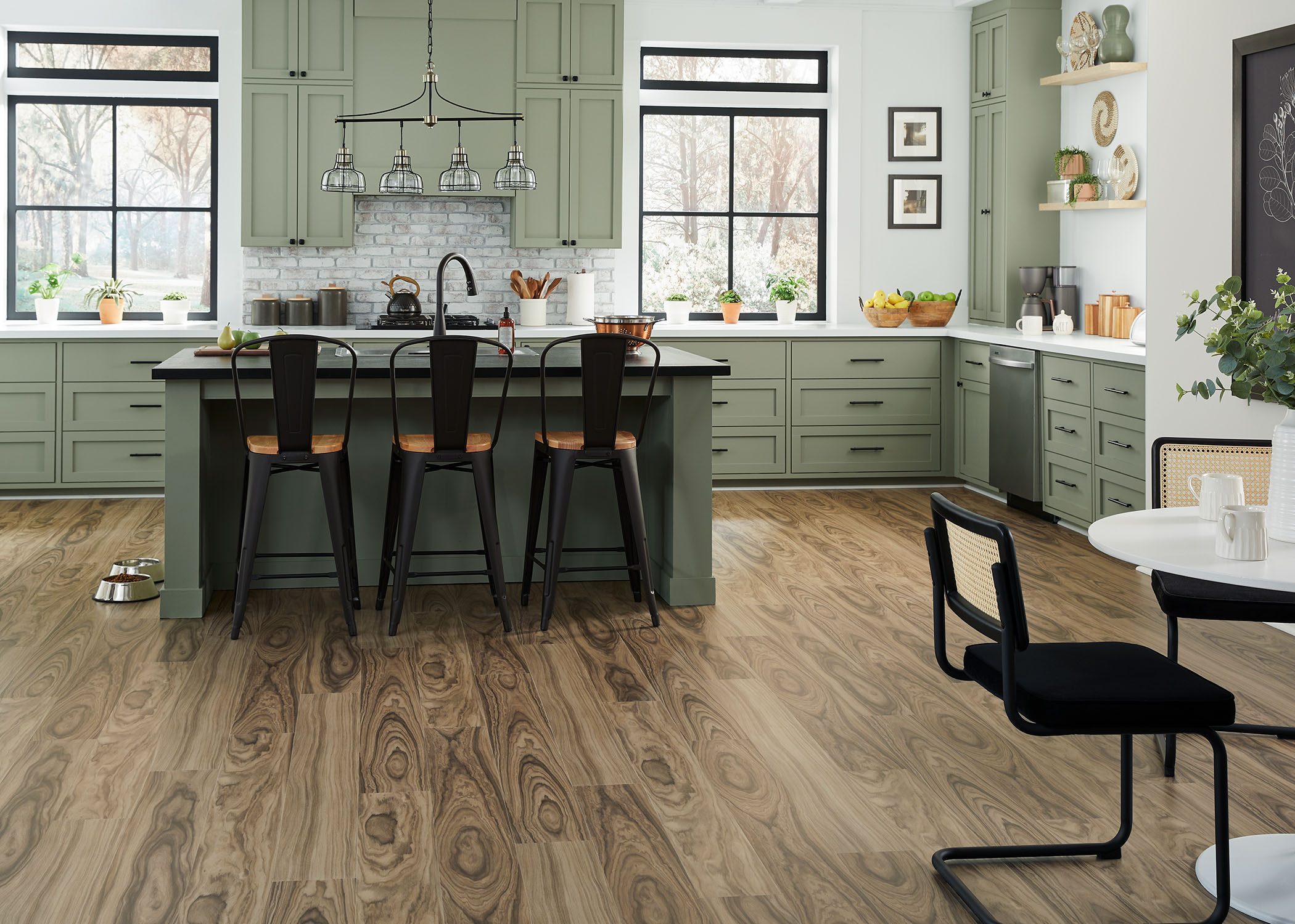 medium brown waterproof rigid vinyl plank floor in kitchen with green cabinets and island plus black metal bar stools with light wood seats and dog bowls on floor and round white dining table and black metal chairs