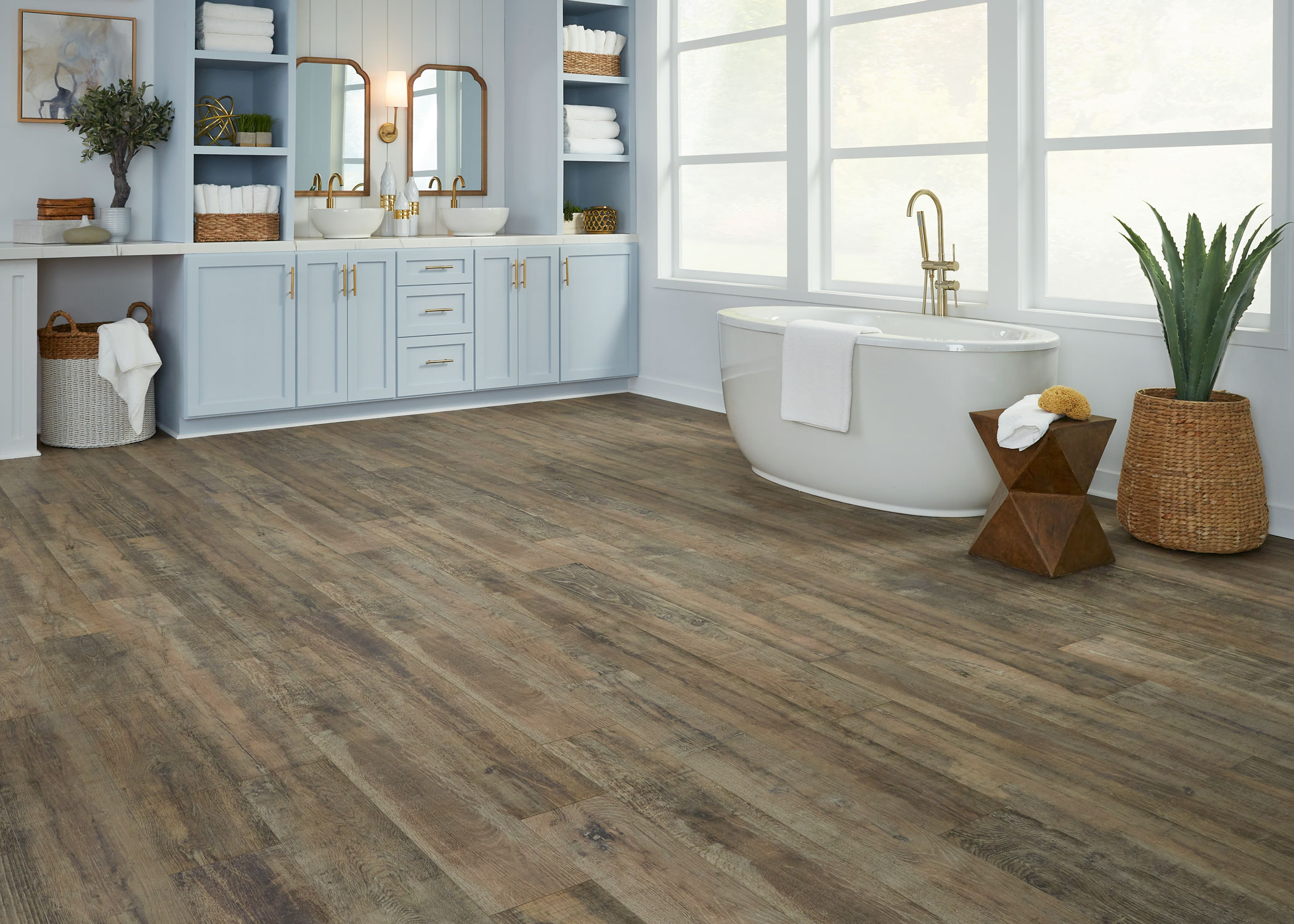 multi toned brown waterproof hybrid resilient flooring in bathroom with pale blue cabinets with dual vessel sinks and white oval freestanding tub