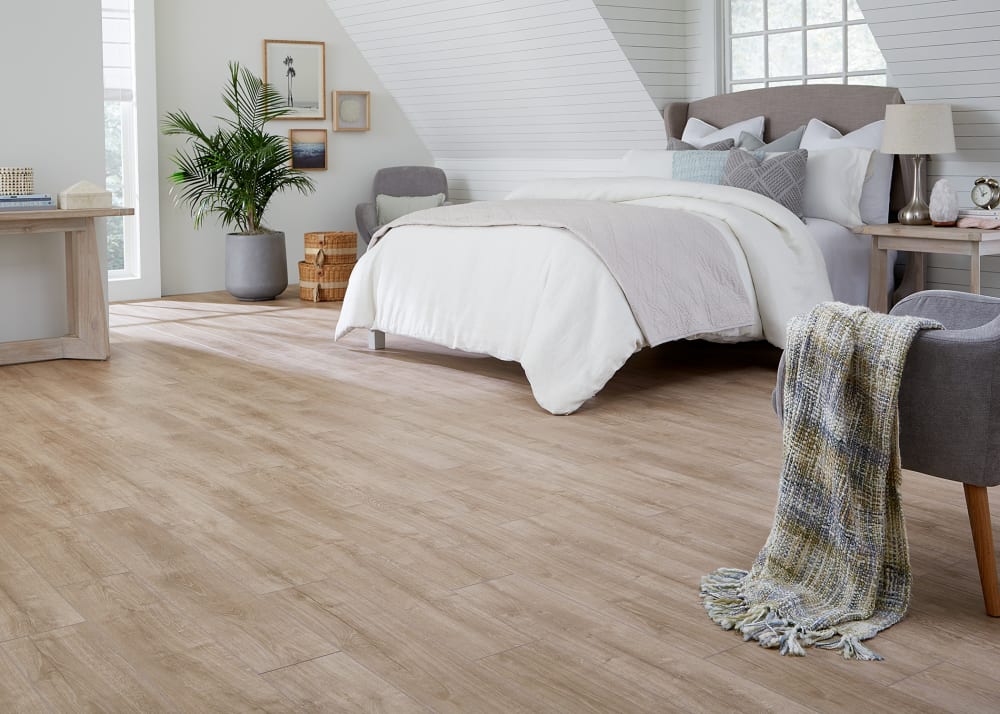 6mm+pad Versailles Oak Rigid Vinyl Plank Flooring in bedroom with white and gray bedding plus gray accent chair with green throw