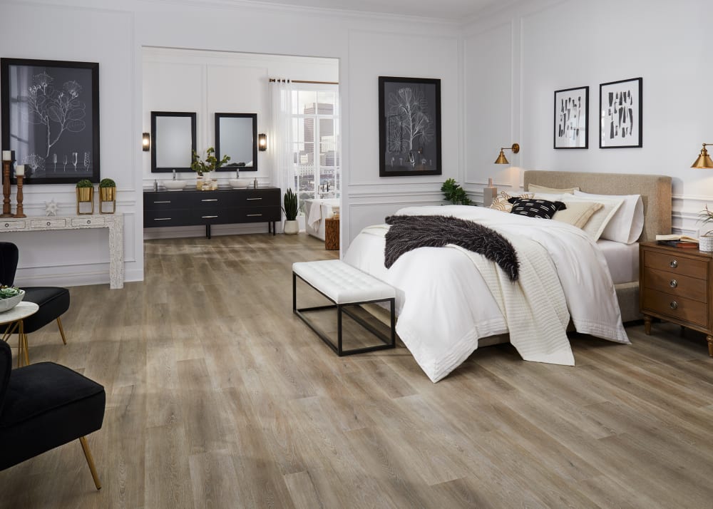 7mm+Pad Chapel Bridge Oak Hybrid Resilient Flooring in primary bedroom with tan upholstered headboard plus white bedding and black and white artwork plus view into bathroom with black dual vanity
