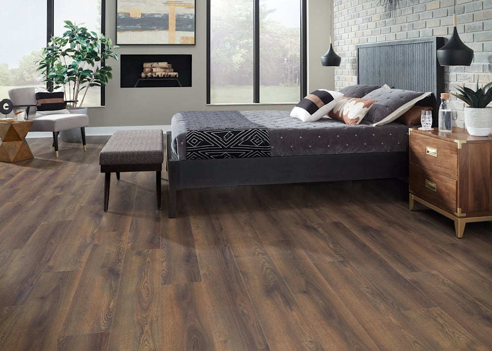 10mm+Pad Barnwood Oak Waterproof Laminate Flooring in bedroom with dark gray velvet bedding and dark brown wood headboard plus brick accent wall and contemporary fireplace plus light gray upholstered accent chair