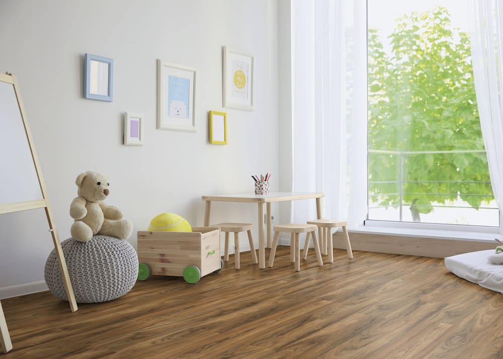 8mm Tradewind Birch Waterproof Laminate Flooring in toddlers playroom with light gray poof and teddy bear sitting on top plus pastel artwork on wall and small table with three stools