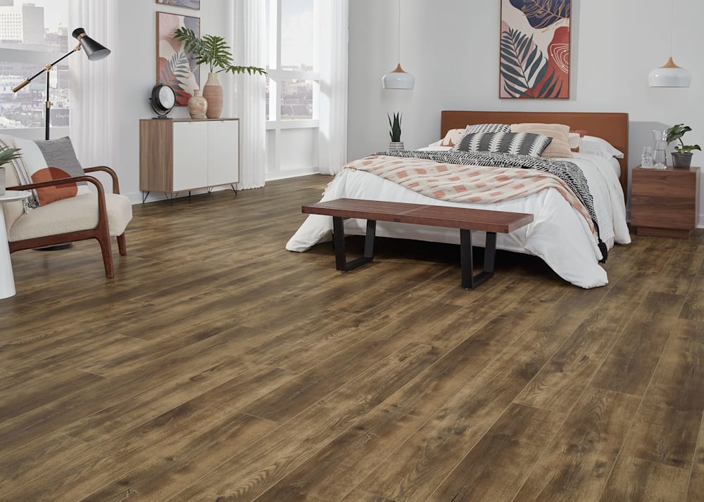 6mm with pad Loma Vista Oak Rigid Vinyl Plank Flooring in bedroom with dark wood headboard and brown and white bedding plus dark wood and metal bench with beige upholstered accent chair and white round side table