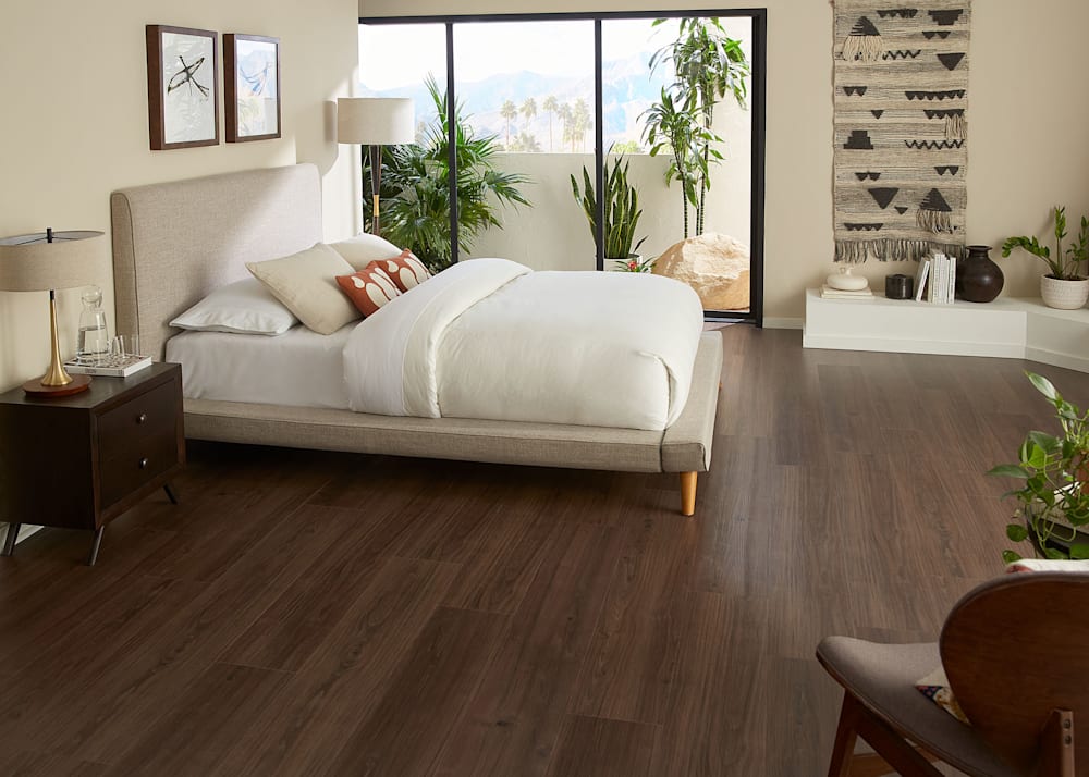 7mm w/Pad Monroe Walnut Rigid Vinyl Plank Flooring in bedroom with beige upholstered platform bed with off white bedding plus pocket glass doors leading to private balcony with desert plants
