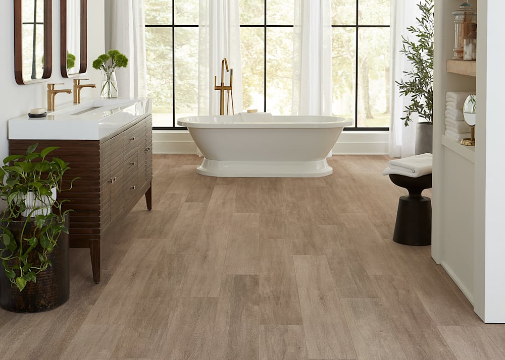 6mm w/Pad Luxembourg Oak Rigid Vinyl Plank Flooring in bathroom with dark brown wood dual vanity with brass fixtures plus freestanding oval tub and oversized potted plant and black round stool with white towel on top