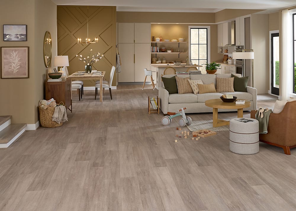 6mm w/Pad Luxembourg Oak Rigid Vinyl Plank Flooring in open concept living, dining and kitchen with beige sofa and caramel accent chair plus wood dining table with cream chairs and white kitchen cabinets with butcher block counters