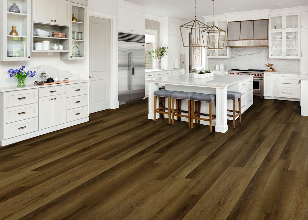 3.2mm Crooksdale Oak Waterproof Rigid Vinyl Plank Flooring in kitchen with white cabinets and oversized white island with gray upholstered wood bar stools and brass fixtures