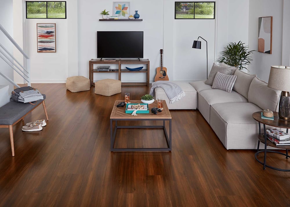 8mm with Pad Revere Oak Waterproof Rigid Vinyl Plank Flooring in lower level family room with beige armless sectional with wood coffee table plus small bench with upholstered seat and flat screen tv on dark wood console